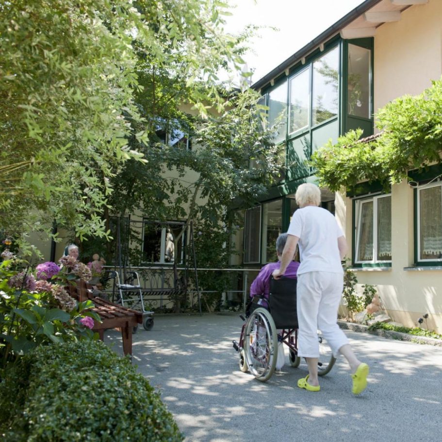 Eine ältere Dame im Rollstuhl wird durch den begrünten Innenhof des Schulhauser Hofs in Iggensbach begleitet.